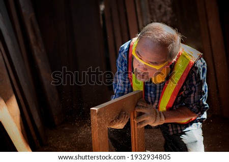 An Asian elderly carpenter in the carpentry shop is checking the workpiece tidy. Wooden window frame That are being repaired and polished with sandpaper to be beautiful Then blow away the dust