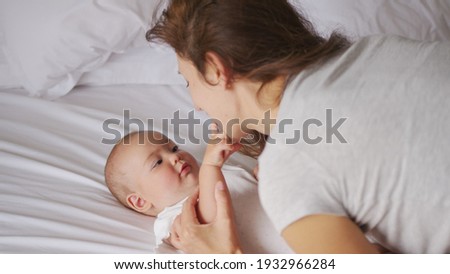 Loving young mommy hugging soothing adorable sweet baby girl lying in bed. Smiling caring mother and cute little infant child girl cuddling in bedroom. Mum and child tender moments.
