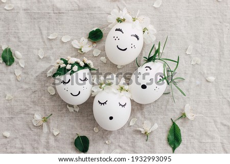 Natural eggs with drawn funny faces in cute floral crowns top view on linen fabric with blooming spring petals and green leaves. Happy Easter! Eco friendly zero waste holiday concept