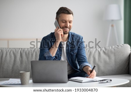 Smiling handsome bearded man having phone conversation from home, using laptop while sitting on couch in living room, talking to business partners, taking notes on notepad, copy space