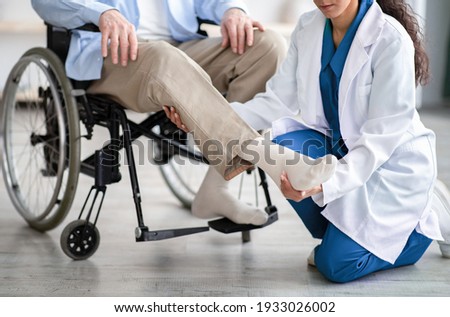 Cropped view of young doctor checking disabled senior patient's leg during medical visit at home. Unrecognizable physiotherapist working with elderly man in wheelchair, closeup Royalty-Free Stock Photo #1933026002
