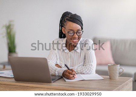 Positive black business woman with stylish glasses manager working from home during COVID-19 pandemic, using modern laptop and taking notes, sitting at workdesk in living room, copy space Royalty-Free Stock Photo #1933029950