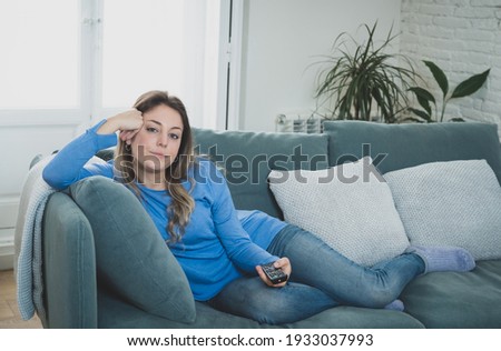 Bored woman watching TV helpless in self Isolation at home during mandatory lockdown due to coronavirus outbreak. Young upset woman on sofa using control remote zapping bored of TV and sedentary life. Royalty-Free Stock Photo #1933037993