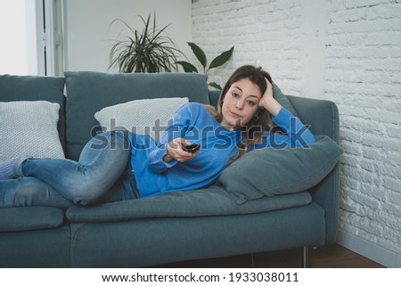 Bored woman watching TV helpless in self Isolation at home during mandatory lockdown due to coronavirus outbreak. Young upset woman on sofa using control remote zapping bored of TV and sedentary life. Royalty-Free Stock Photo #1933038011