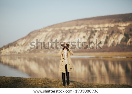 Young stylish girl in hat and trendy trench coat takes photo on retro camera with mountain and river on background. 