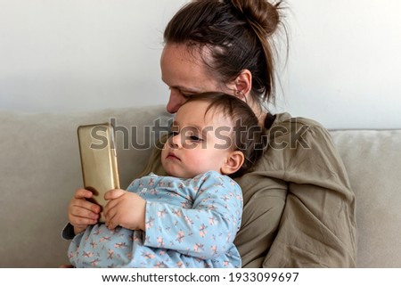 Mother and daughter looking at mobile phone at home. Happy pretty mommy cuddling baby daughter, while looking at smartphone at home. Loving young mom embracing little child, using mobile apps together