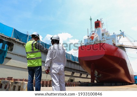 Engineering Workers planning hand holding blue print of the commercial ship standing on floating dry dock yard, recondition, repairing, painting overhaul in dock yard in shipyard Royalty-Free Stock Photo #1933100846