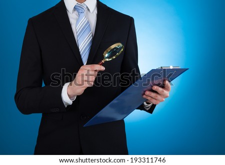 Midsection of businessman examining document on clipboard with magnifying glass against blue background