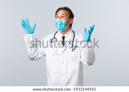 Covid-19, preventing virus, healthcare workers and vaccination concept. Delighted and relieved male doctor in medical mask, gloves, raising hands up happy, looking top left corner pleased