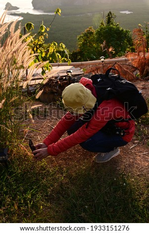 A Tourists wear bright red duck feathers. Wear jeans, a black backpack and a yellow hat, sit and use your mobile phone to take beautiful pictures of Golden grass flower as the sunrise on the mountain.