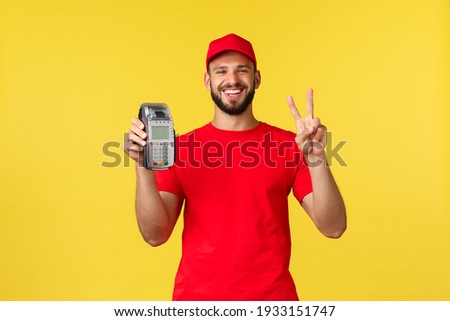 Contactless payment, online delivery, internet shopping and parcel tracking concept. Funny friendly courier in red uniform cap and t-shirt, employee make peace sign and show POS terminal