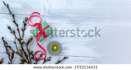 Green gift box and candle. Willow branches on a gray-blue background. A gift with a red ribbon.