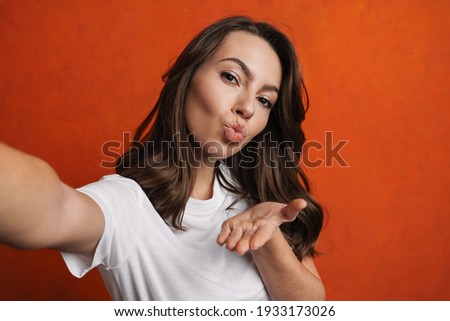 Happy beautiful girl blowing air kiss while taking selfie photo isolated over orange wall
