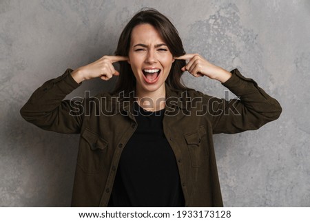 Nervous beautiful girl screaming and plugging her ears isolated over grey wall