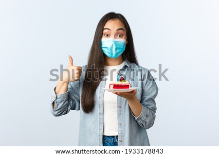 Social distancing lifestyle, covid-19 pandemic, celebrating holidays during coronavirus concept. Happy smiling asian birthday girl in medical mask, show bday cake and thumb-up pleased