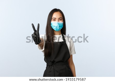 Covid-19, social distancing, small coffee shop business and preventing virus concept. Smiling cute asian female barista, restaurant or cafe worker in medical mask and gloves show v-sign, peace