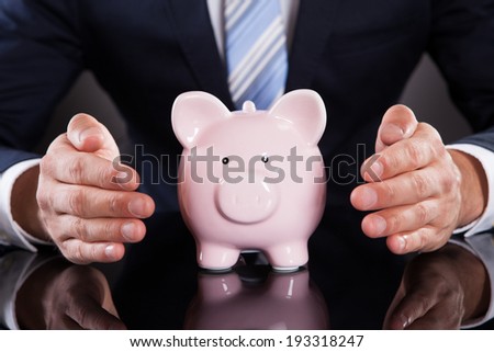 Midsection of businessman protecting piggybank with cupped hands on desk against black background