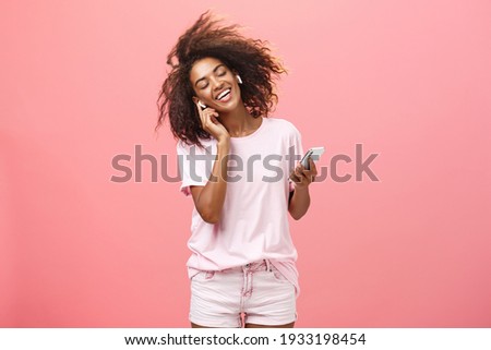 Getting rid of worried with good tunes in ears. Pleased attractive young african woman in trendy outfit listening music in wireless earphones touching earbud smiling dancing and holding smartphone Royalty-Free Stock Photo #1933198454