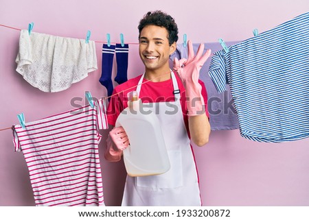 Handsome hispanic man doing laundry holding detergent bottle doing ok sign with fingers, smiling friendly gesturing excellent symbol 