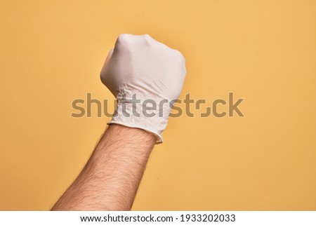 Hand of caucasian young man with medical glove over isolated yellow background doing protest and revolution gesture, fist expressing force and power