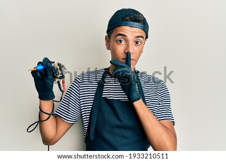 Young handsome african american man tattoo artist wearing professional uniform and gloves holding tattooer machine asking to be quiet with finger on lips. silence and secret concept.  Royalty-Free Stock Photo #1933210511