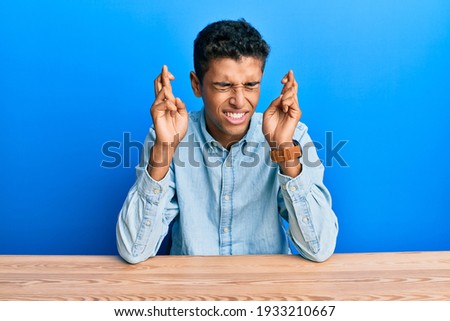 Young handsome african american man wearing casual clothes sitting on the table gesturing finger crossed smiling with hope and eyes closed. luck and superstitious concept. 