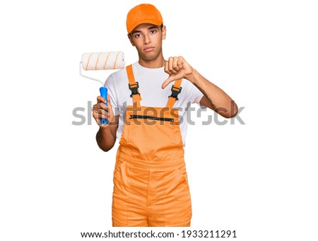 Young handsome african american man wearing cap and painter clothes holding painting roll with angry face, negative sign showing dislike with thumbs down, rejection concept 