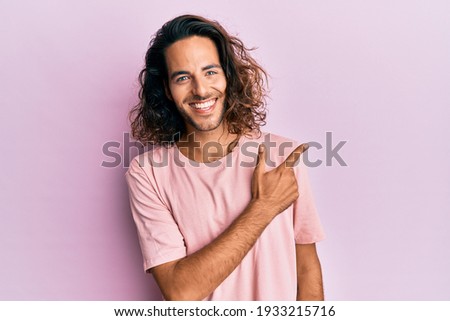 Young handsome man with long hair wearing casual clothes smiling cheerful pointing with hand and finger up to the side 