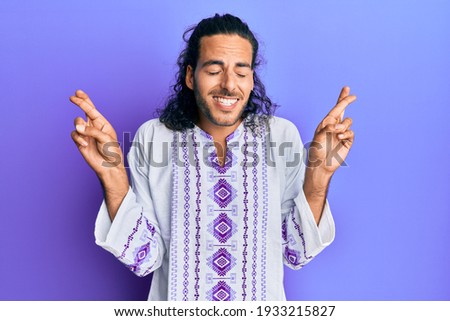 Young handsome man with long hair wearing bohemian and hippie shirt gesturing finger crossed smiling with hope and eyes closed. luck and superstitious concept. 