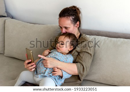 Caucasian mother and child playing game on the smartphone at home. Mother with baby daughter sitting on sofa at home looking at mobile. Mom and baby with a phone at home watching cartoons and playing.