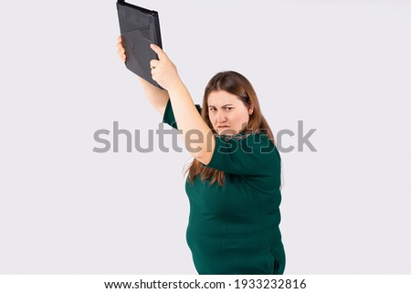 Young blonde overwight businesswoman in green dress over gray background, frustrated large woman holding pc computer up. Indignant girl, female character hitting a laptop. Mad office worker concept