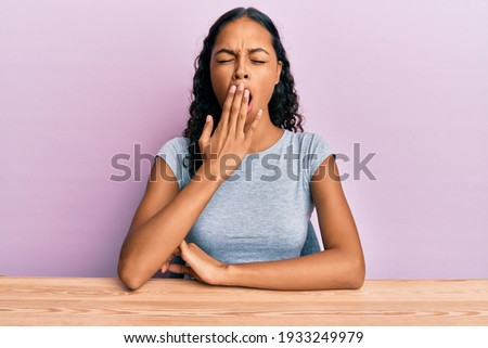 Young african american girl wearing casual clothes sitting on the table bored yawning tired covering mouth with hand. restless and sleepiness. 