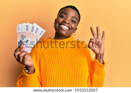 Young african american woman holding icelandic krona banknotes doing ok sign with fingers, smiling friendly gesturing excellent symbol  Royalty-Free Stock Photo #1933252007