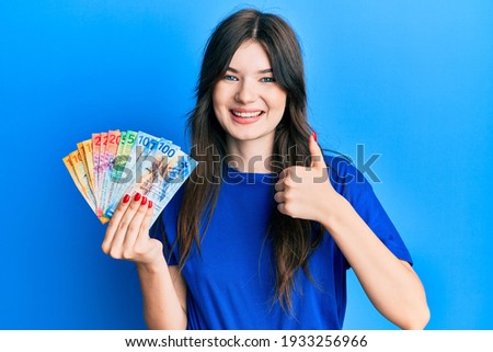 Young beautiful caucasian girl holding swiss franc banknotes smiling happy and positive, thumb up doing excellent and approval sign 