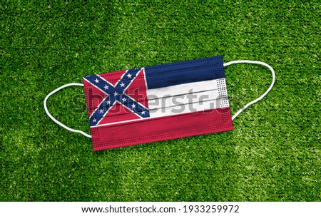 Flag Mississippi placed on a medical mask lies on the green grass. Covid-19 pandemic concept