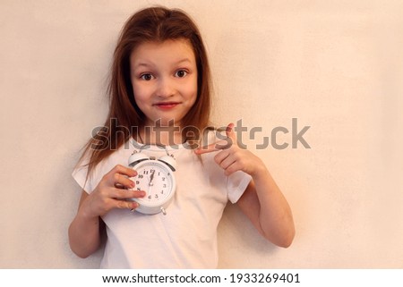 An eight-year-old girl in a white T-shirt with untidy hair in a good mood, with a watch in her hands, points at it with the other hand - the concept of the morning rise and the beginning of the day