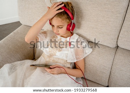 Little girl sitting on the sofa in living room using headphone and smartphone, watching cartoons or listening music. Girl dressed in a beautiful white tulle fancy dress. High quality photo