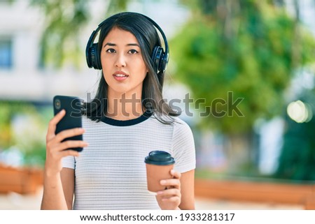 Young hispanic woman drinking coffee and using smartphone and headphones at the city.
