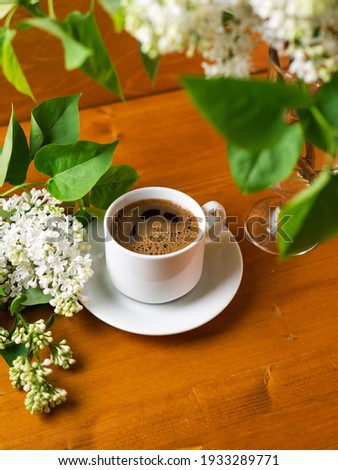 White cup of fragrant hot espresso coffee on a wooden table, bouquet of white lilac, copy space