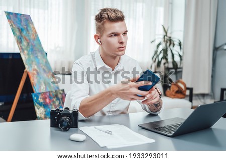 Male designer with photo camera and laptop doing his job in modern office. Tattooed guy dressed in white shirt sits at table using his phone.