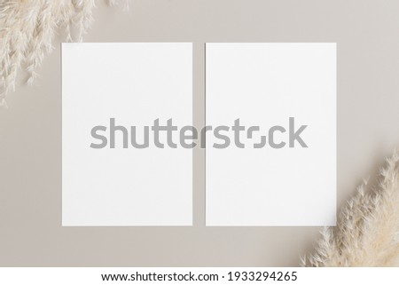 Two white invitation cards mockup with a pampas decoration on a beige table. 5x7 ratio, similar to A6, A5.