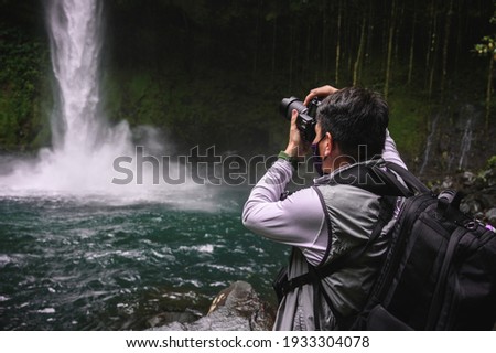 Handsome male photographer with a waterfall as background. Man photographing La Fortuna Waterfall at Costa Rica.