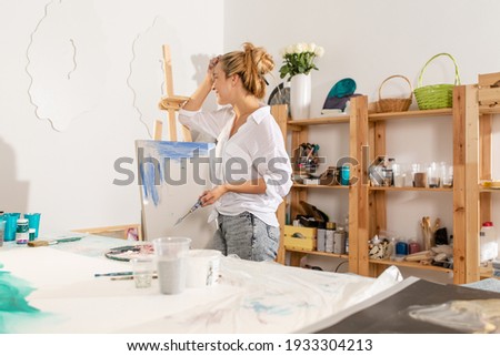 An artist standing in front of an easel painting a picture with paints standing in a creative art studio 