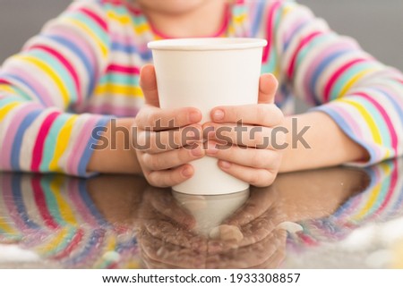  CUTE Beautiful little blonde girl looks at the camera and smiles while sitting in a cafe and sipping a milkshake drink from a white paper cup mug horizontal photo