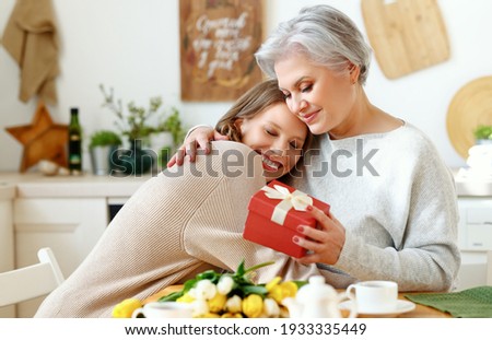 Cheerful young woman hugging happy elderly female with closed eyes and giving flowers and present box on Mother Day