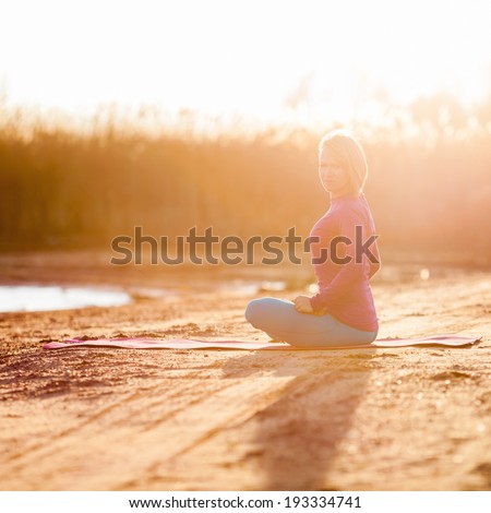 Young woman doing yoga near lake outdoors, meditation. Sport fitness and exercising in nature, autumn sunset