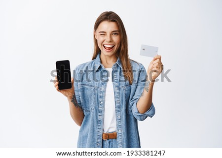 Online shopping. Cheeky young woman winking at you, recommending mobile banking app, showing empty smartphone screen and plastic credit card, white background