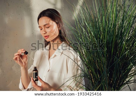Woman hands holding massage or cosmetics oil bottle for applying drops to skin of face. Female hold oil on old wall background with shade from foliage. Concept of healthy lifestyle and self care Royalty-Free Stock Photo #1933425416