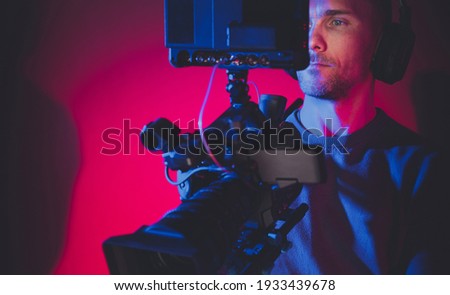Video Production Stage Dark Red and Blue Illuminated and Film Making Professional Digital Camera Operator at Work. Caucasian Men in His 40s.
