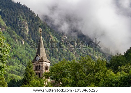 bell tower with a clock in a mountain village. Clouds cover a hillside in the background, in Arties, Valle de Aran Lleida, Spain Royalty-Free Stock Photo #1933466225